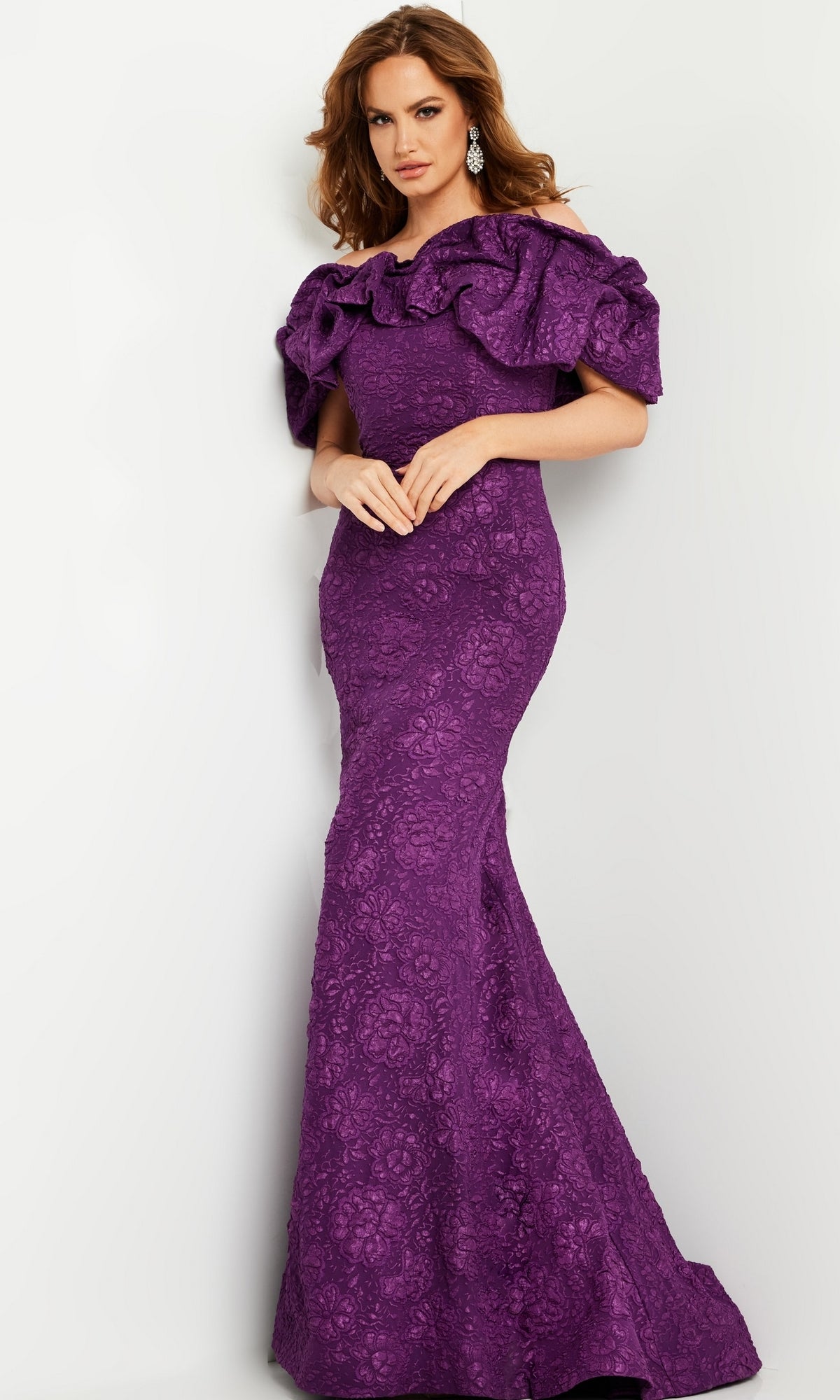 Woman wearing the Jovani Long Brocade Mermaid Prom Dress 23847 featuring a floor-length, purple design with an off-the-shoulder ruffled neckline and floral lace texture, standing against a plain light background.